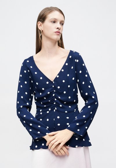 Μπλε σκούρο τοπ με μακριά μανίκια και λευκές πουά, σχεδιασμένο με V-λαιμόκοψη, ρίγες στο μπροστινό μέρος και φαρδύ στρίφωμα. Μαλακή υφή, ελαφρύ ύφασμα.