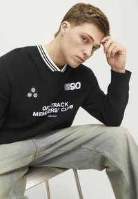 Sort tekstureret sweatshirt med hvid stribet krave, udstyret med logo patches og broderet tekst. Modellen har lysegrå denim jeans på.