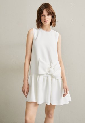 Vestido branco sem mangas feito de tecido macio, com um corpete justo, saia rodada e um grande laço destacado na cintura.