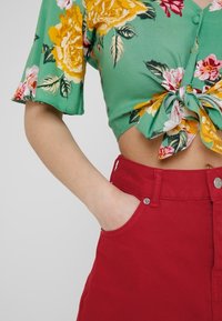 Blusa cropped floreale verde con rose gialle e rosa, maniche corte e nodo davanti. Abbinata a pantaloni rossi a vita alta.