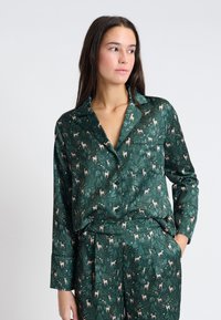 Conjunto de pijama verde de satén con patrones de ciervos y bosques, que incluye una camisa con cuello y pantalones a juego, ambos con una textura suave.