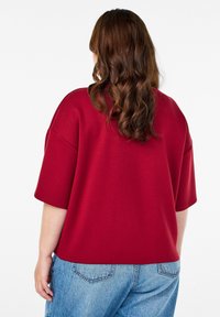 Bordový voľný sveter s krátkymi rukávmi, hladká textura a ležérny strih, v kombinácii s jasne modrými džínami. Zobrazený zadný pohľad.