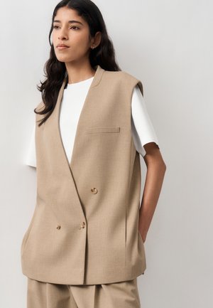 Kvinne med langt mørkt hår som har på seg en ermeløs beige blazer over en hvit kortermet skjorte, stående foran en ensfarget lys bakgrunn.