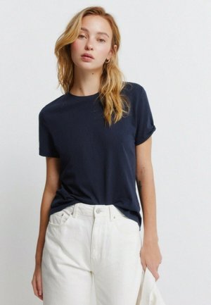 Młoda kobieta z rozpuszczonym blond włosami, ubrana w granatowy t-shirt z krótkim rękawem i białe spodnie z wysokim stanem, trzymająca biały materiał.