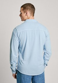 Camisa de manga larga azul claro con una textura suave, puños con botones y un dobladillo curvado, que presenta un cuello clásico y un diseño sencillo.