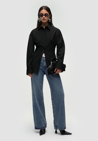 Sort oversized skjorte med knapper, brede blå denimjeans, spidse sorte hæle, sort håndtaske med tekstureret finish og mørke solbriller.