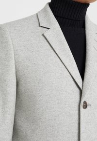 Blazer de lana gris claro con solapa entallada, cierre de un solo botón y textura sutil, combinado con un suéter de cuello alto negro.