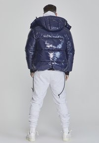 Navy puffer jacket with a glossy finish and an attached hood, featuring horizontal quilted sections. Paired with light gray sweatpants.