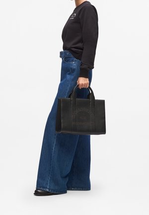 Femme en sweat-shirt noir et jean coupe large tenant un sac fourre-tout en cuir noir avec logo embossé et doubles poignées.