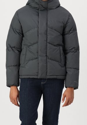 Person iført en sort dunjakke med diagonale syninger og sidelommer, kombineret med mørkeblå jeans, stående mod en ensfarvet baggrund.