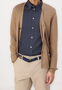 Brown knit cardigan over a navy button-up shirt, paired with beige pants and a braided navy belt. Close-up on fabric texture and button details.