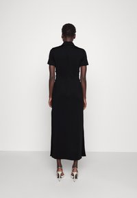 Mustan, lyhyet hihat omaava maksimekko, jossa on istuva siluetti, sivurei'itykset ja korkea kaulus. Mallilla on päällään hopeiset korokepohja sandaalit.