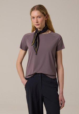 Femme aux longs cheveux blonds portant un haut mauve à manches courtes, un pantalon noir et un foulard noir et or autour du cou, debout avec une main dans la poche.