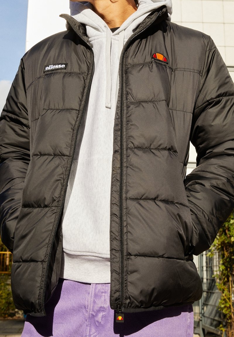 Czarna kurtka puchowa z pikowanym wzorem, zapinana na zamek błyskawiczny, z logo. Noszona na szarym hoodie, w połączeniu z fioletowymi spodniami.