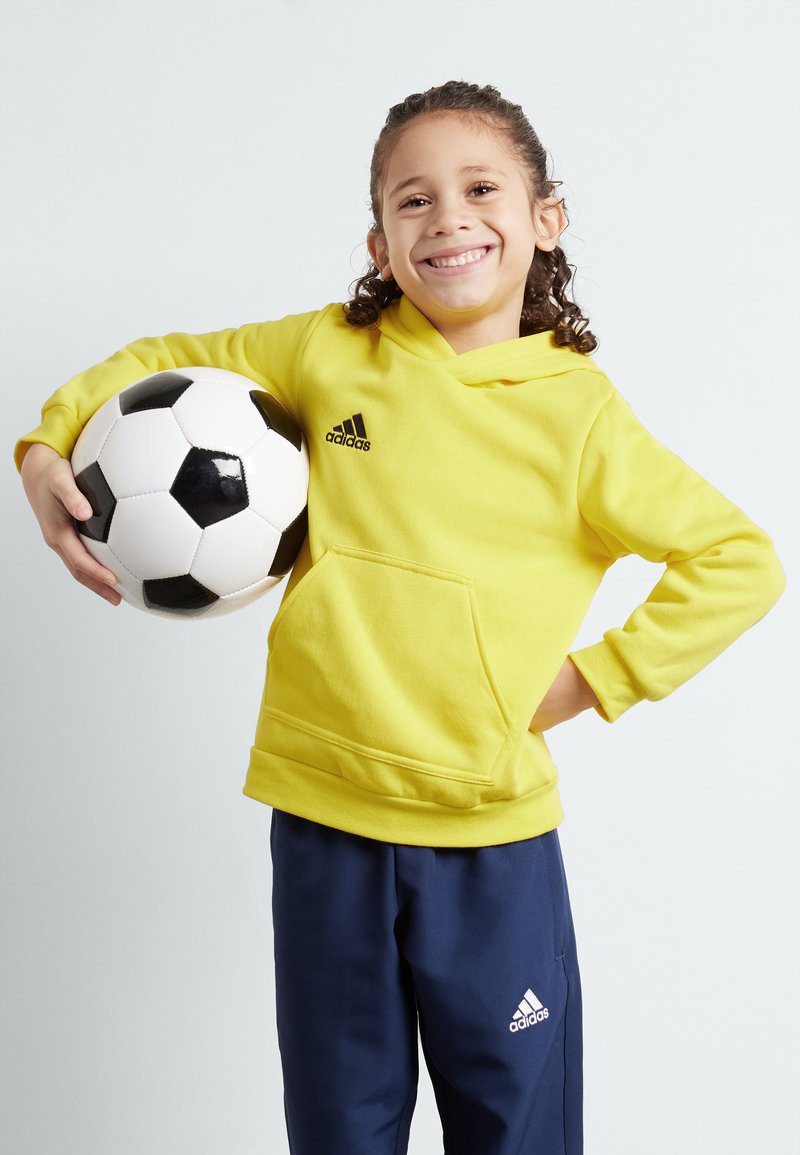 Felpa gialla con cappuccio e tasca frontale, logo Adidas nero, abbinata a pantaloni blu navy con logo Adidas bianco, mentre tiene un pallone da calcio bianco e nero.