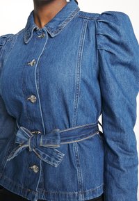 Džínová bunda s nafouknutými rukávy, světle modré barvy, pěti kovovými knoflíky a zavazovací látkový pásek v pase. Obsahuje viditelné švy.