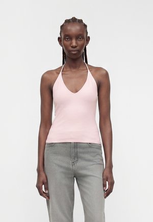 Femme avec des tresses portant un haut dos nu rose clair et un jean gris, debout sur un fond blanc uni.