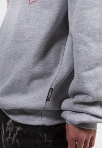Close-up de una persona que lleva una sudadera gris con una etiqueta negra que dice "WASTED" y pantalones negros con un patrón de salpicaduras de pintura.