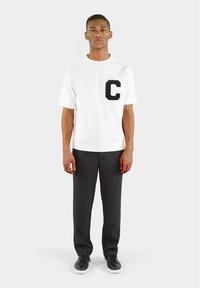 Hvit t-skjorte med en svart "C"-lapp på venstre bryst, kombinert med grå stripete bukser og svarte sneakers. Enkel, uformell design.