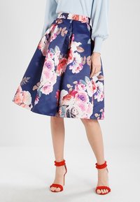 Kvinna i en marinblå A-linje kjol med stora rosa och vita blommönster, kombinerad med röda högklackade sandaler och en ljusblå tröja.