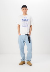 T-shirt alb cu imprimeu albastru "Red Bull Racing", blugi din denim albastru deschis și adidași bej. Bandană neagră legată la talie. Design simplu.