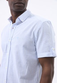Camisa de botones de manga corta en rayas azul claro y blanca, con cuello botón y un logo en el pecho. Fabricada con tejido ligero.