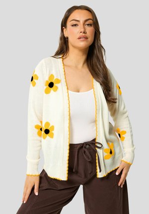 Femme portant un cardigan blanc avec des motifs de fleurs jaunes, un débardeur blanc et un pantalon marron foncé avec cordon, debout devant un fond uni.