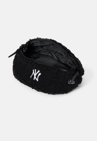 Svart fuskpäls-påse med dragkedjestängning, med en broderad vit logotyp på framsidan och en slät svart foder inuti.