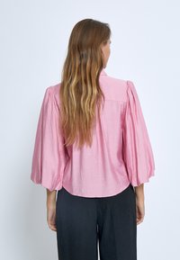 Blusa di lino rosa con maniche a sbuffo e orlo arrotondato. La parte posteriore presenta una texture sottile ed è abbinata a pantaloni neri.