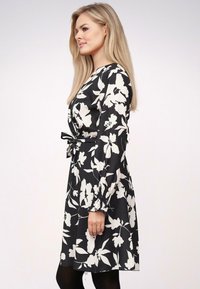 Vestido negro con patrón floral blanco, de manga larga y con puños elásticos. Cinturón anudado en la cintura, hasta la rodilla, con tela fluida.
