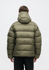 Person facing away wearing an olive green puffer jacket with hood and black pants with red accents against a plain white background.