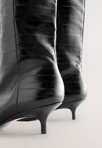 Bottines en cuir noir brillant avec petits talons kitten, présentant des détails de couture horizontale texturée sur surface lisse.