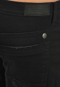 Melns denims ar ādas jostas etiķeti no B.LEND. Sabojātu kabatu malas un sudraba tonis metāla detaļas.