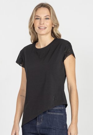 Kobieta ubrana w czarną asymetryczną bluzkę z krótkim rękawem i przezroczystymi rękawami oraz ciemnoniebieskie dżinsy, uśmiechająca się na tle białego tła.