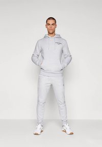 Σετ γκρι φούτερ με κουκούλα και παντελόνι φόρμας από βαμβάκι, με μπροστινή τσέπη και λογότυπο στο στήθος. Συνδυάζεται με λευκά αθλητικά παπούτσια.