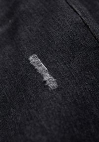 Tissu noir à texture rugueuse présentant un détail de couture blanche irrégulière et proéminente. La surface semble légèrement usée.