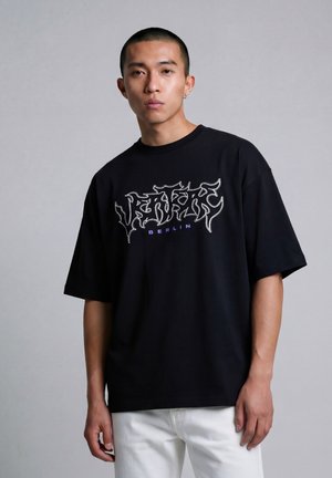 Jonge man met kortgeknipt haar, draagt een zwart oversized T-shirt met graffiti-stijl tekst en "BERLIN" in paars, gecombineerd met witte broeken.