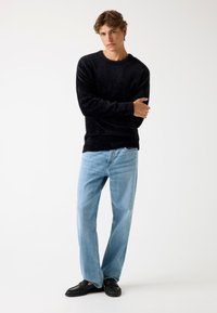 Zwarte fuzzy trui, lichtblauwe rechte jeans en zwarte platte schoenen. De trui heeft een ronde halslijn en een gestructureerde stof.
