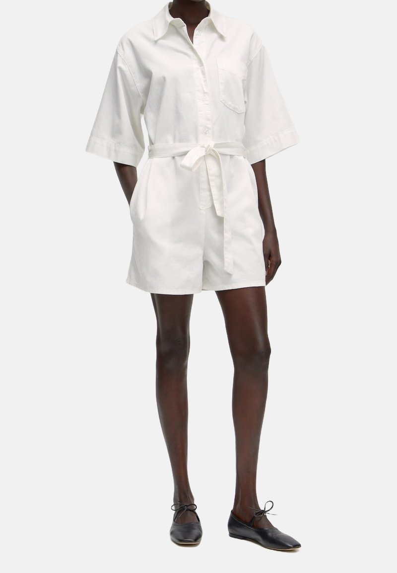 Femme portant une combinaison-short blanche à manches courtes ceinturée avec des poches avant et des ballerines noires, debout devant un fond uni.