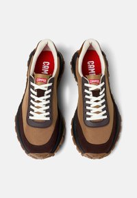 Sneakers marroni e beige con una tomaia in tessuto texturizzato, lacci bianchi e una suola robusta nera e marrone. Logo rosso visibile all'interno.