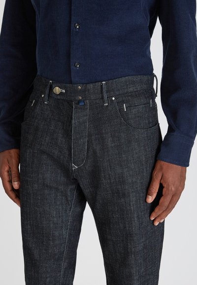 Mann trägt ein dunkelblaues Kordhemd und dunkelgraue Jeans, Hände entspannt an den Seiten, Oberkörper sichtbar.