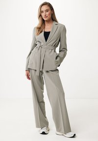 Blazer grigio chiaro su misura con cintura a cravatta, pantaloni a gamba larga abbinati e una texture liscia; abbinato a sneakers bianche.