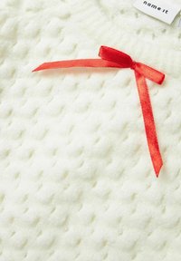 Vit texturerad tyg med ett upphöjt mönster, prydd med en liten röd sidentofs detalj. Märkesetikett synlig vid ovankanten.