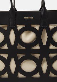 Sac fourre-tout noir avec un design géométrique découpé au laser, révélant un intérieur en toile beige. Il dispose de doubles anses en cuir noir et d'un détail de logo doré.
