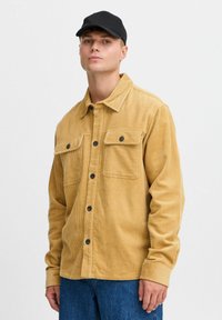 Katoenen shirtjack in mosterdgeel, met een knoopsluiting aan de voorkant, twee borstzakken en zwarte knopen. Model draagt een zwarte pet.