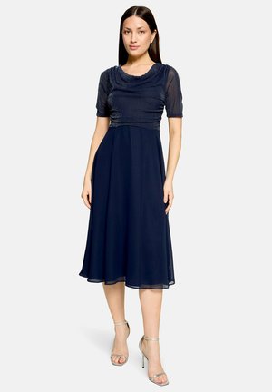 Femme portant une robe midi bleu marine avec des manches courtes transparentes et des sandales argentées à talons hauts, debout devant un fond clair uni.