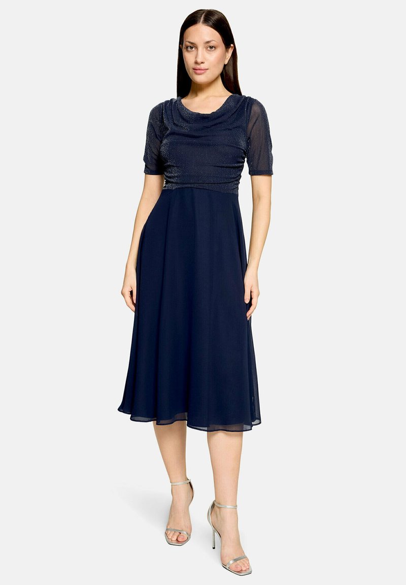 Femme portant une robe midi bleu marine avec des manches courtes transparentes et des sandales argentées à talons hauts, debout devant un fond clair uni.