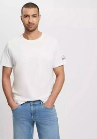 Biały bawełnowy T-shirt z okrągłym dekoltem i krótkimi rękawami. Ma małe paskowane logo na lewym rękawie. Nosi się go z jasnoniebieskimi dżinsami.