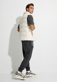 Weiße, wattierte Weste mit Kapuze, ärmelloses Design, getragen über einem dunkelgrauen Shirt. Ein Paar tapered, dunkle Jogginghosen und gemusterte Turnschuhe.