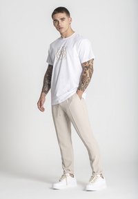 Mladý muž s tetovanými rukami, oblečený v bielom tričku s grafickým motívom, béžových nohaviciach a bielych teniskách, stojí pred jednoduchým svetlým pozadím.